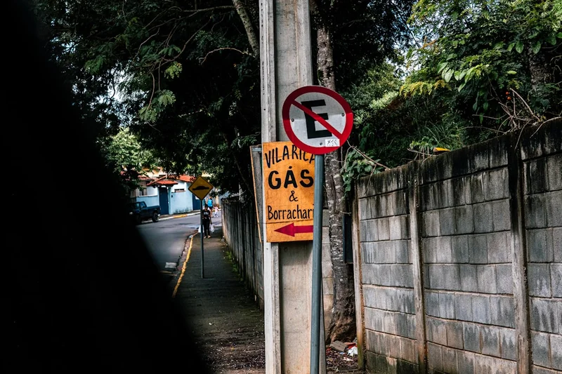 A woman walks down a sidewalk past signs for a gas station and tire shop, with a no parking sign overhead.